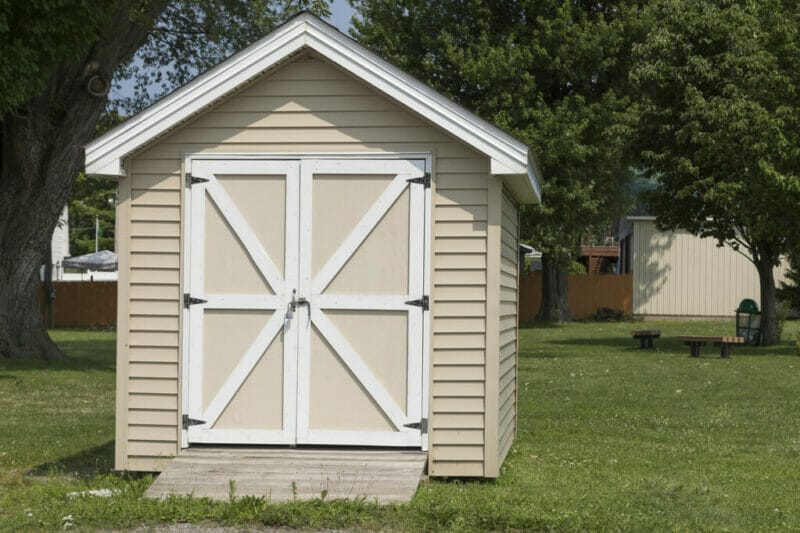 Small beige shed with ramp in backyard. Cute size for lawnmower storage.
